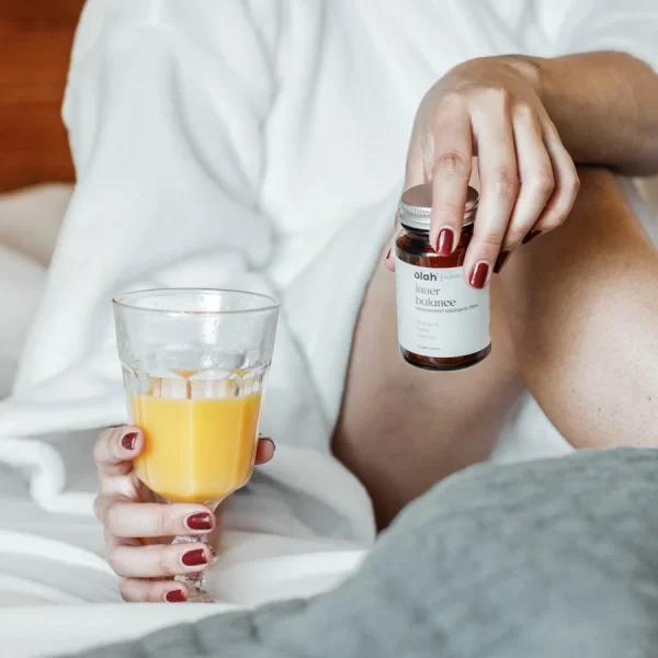 A person with red painted nails and white bathrobe sits in bed holding a bottle of Inner Balance hemp powder capsules and an orange drink in a glass chalice